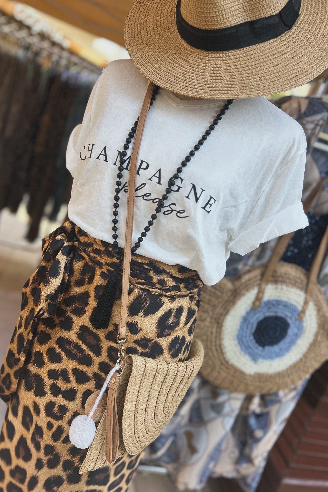 Person wearing a white t-shirt with 'Champagne Please' text, leopard print wrap skirt, straw hat, and carrying a tan clutch bag.